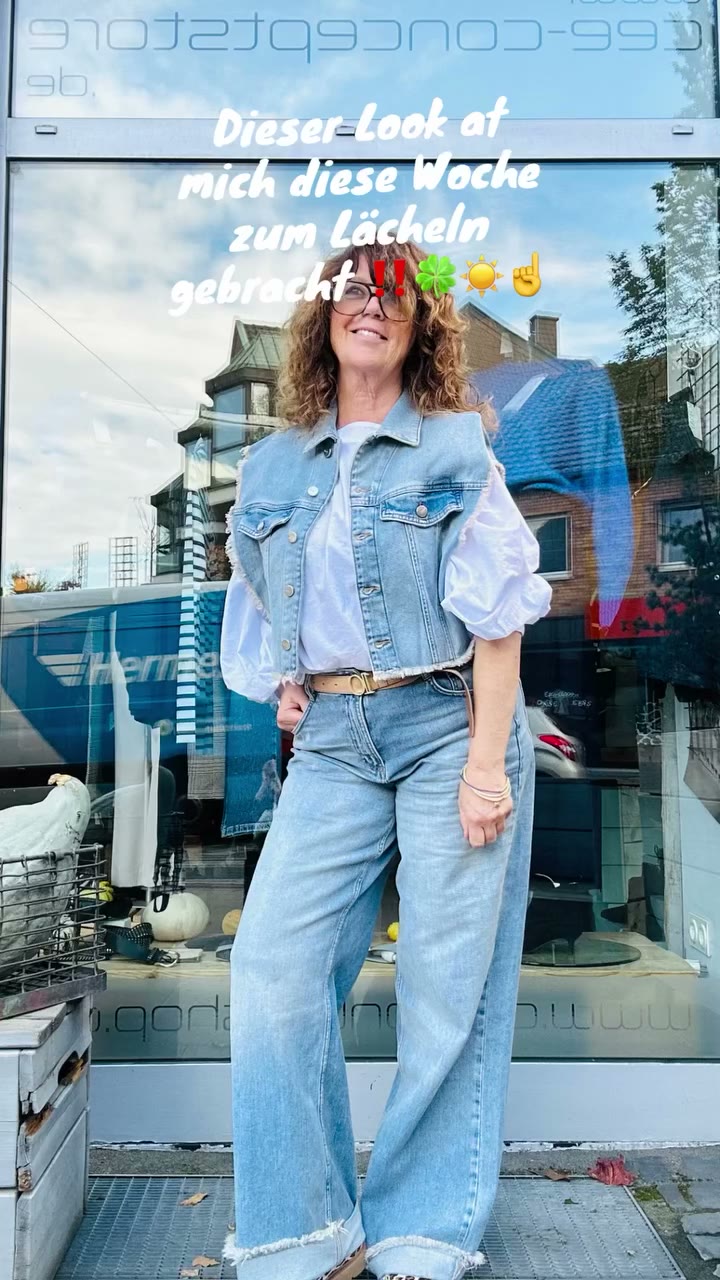 CEE im Denim-Look mit Jeansweste, weißer Bluse mit Puffärmeln und weiter heller Jeans mit Fransensaum vor dem Store.