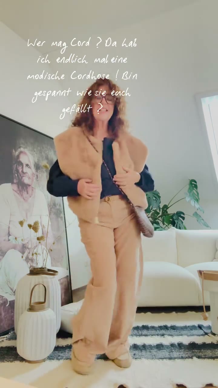 CEE in weiter camelfarbener Cordhose mit dunkelblauem Pullover und beiger Teddy-Weste im hellen Wohnraum.