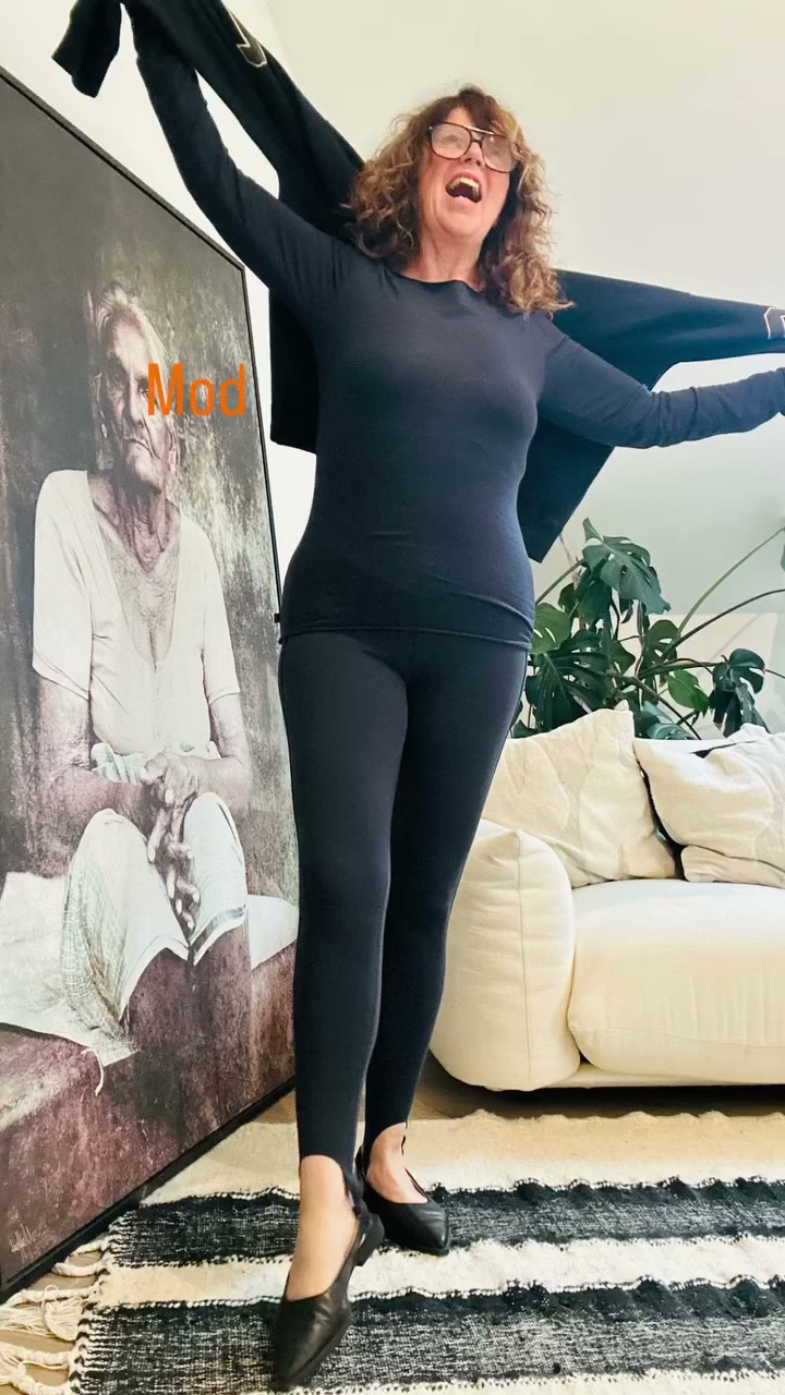 CEE ganz in Schwarz mit Legging, Longsleeve und flachen Slingback-Schuhen, lachend mit Jacke in der Hand.